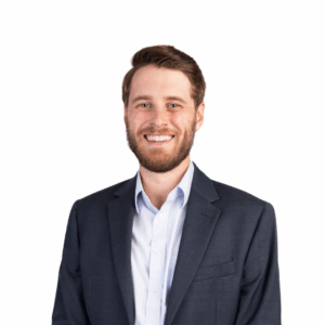 A smiling man with short brown hair and a beard, wearing a dark suit jacket and a light blue shirt, stands against a plain white background.