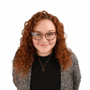 A woman with long curly red hair and glasses smiles at the camera. She is wearing a black top, a gray patterned jacket, and a triangular pendant necklace, standing against a plain white background.
