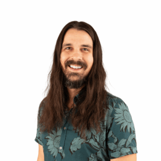 A man with long brown hair and a beard smiles at the camera. He is wearing a green shirt with a sunflower pattern, standing against a plain white background.
