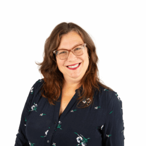 A smiling woman with long brown hair and glasses, wearing a navy blue blouse with a floral pattern, stands against a plain white background.