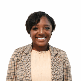 A woman with short, wavy hair smiles at the camera. She is wearing a beige blouse and a plaid blazer. The background is plain white.