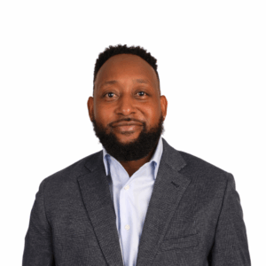 A man with short curly hair and a beard, wearing a blue checked blazer over a light blue shirt, smiling slightly, standing against a plain white background.