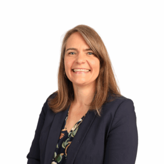A woman with straight, light brown hair smiles at the camera. She is wearing a navy blazer over a patterned blouse and is posed against a plain white background.