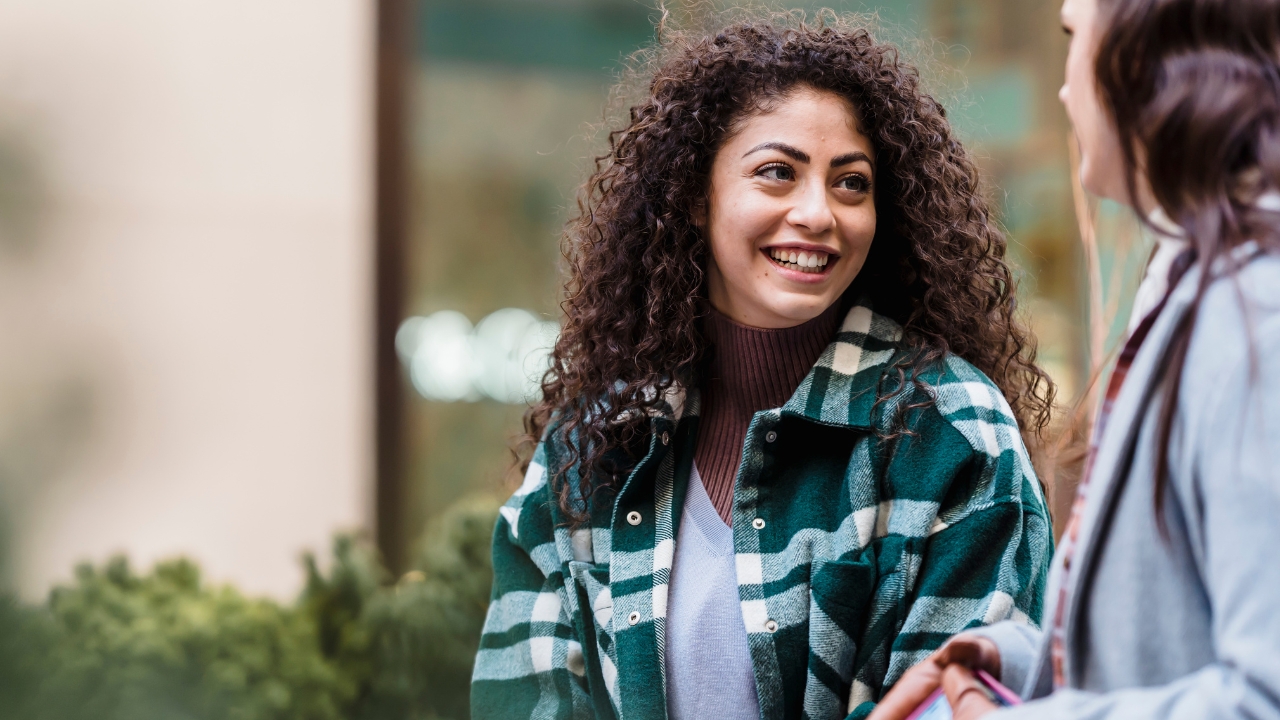Two people smiling and talking outdoors. One person has curly hair and is wearing a green plaid jacket and turtleneck. The other person has straight hair and is wearing a light coat. Greenery and blurred background visible.