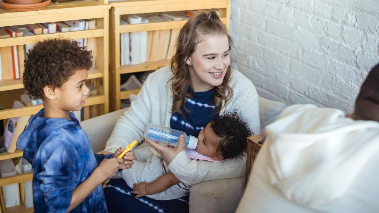 A woman sitting in a chair smiles while feeding a baby with a bottle. A child stands nearby holding a yellow toothbrush. Bookshelves filled with books are in the background.