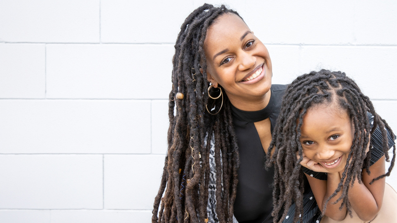 A smiling woman with long dreadlocks embraces a smiling child with braided hair. They are standing against a white brick wall and appear joyful and content.