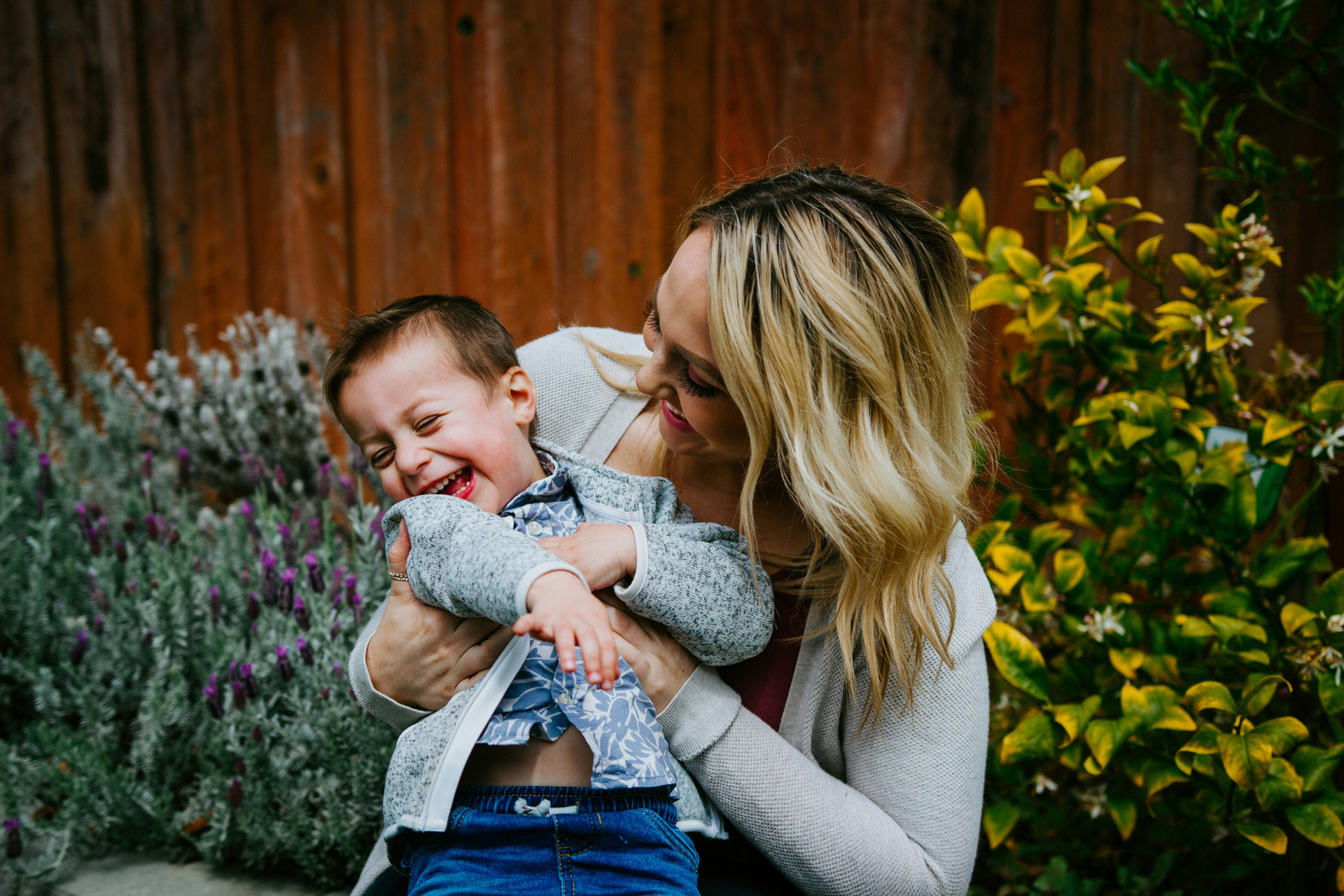 A woman with long blonde hair holds a young boy who is laughing joyfully. They are sitting outdoors near a wooden fence, surrounded by flowering plants. Both are wearing casual clothing, and the scene conveys warmth and happiness.