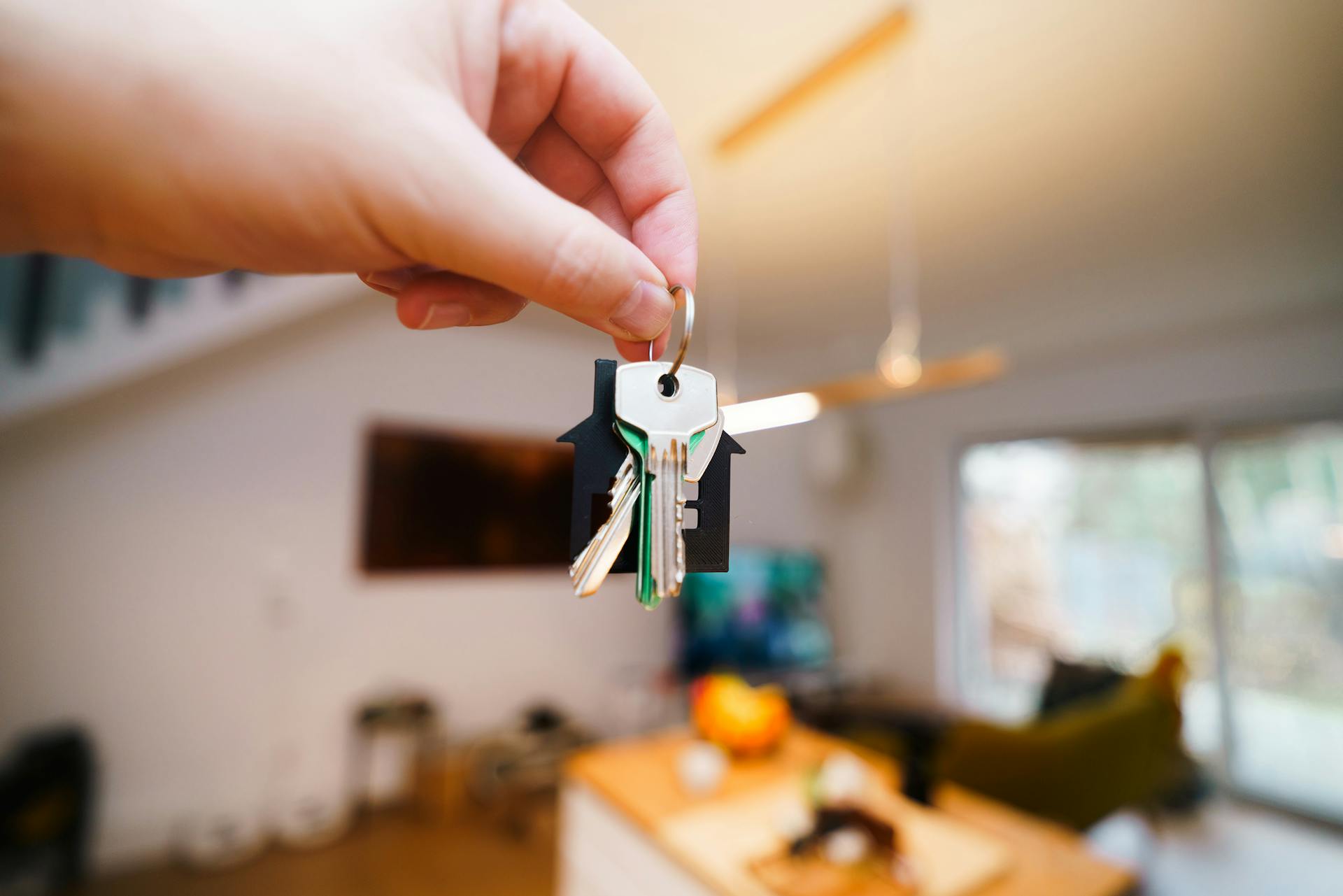 A hand holding a set of keys with a house-shaped keychain in focus. The background shows a blurred indoor living space with modern decor, including a ceiling fan and a large sliding glass door.