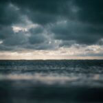 Dark clouds hover above a tranquil, expansive sea, creating a moody and atmospheric scene with a hint of light peeking through the sky.