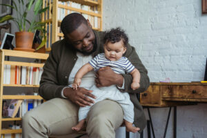 A man sits in a cozy room, holding a baby on his lap. The man smiles affectionately at the baby, who is wearing a striped shirt and light pants. Books and a plant are on shelves in the background.