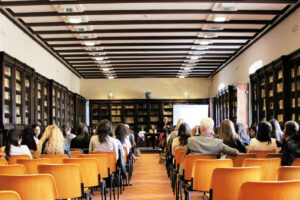 A conference room with rows of orange chairs facing a panel of speakers at the front. The room has wooden bookshelves filled with books along the walls, and several people are seated, attending the event.