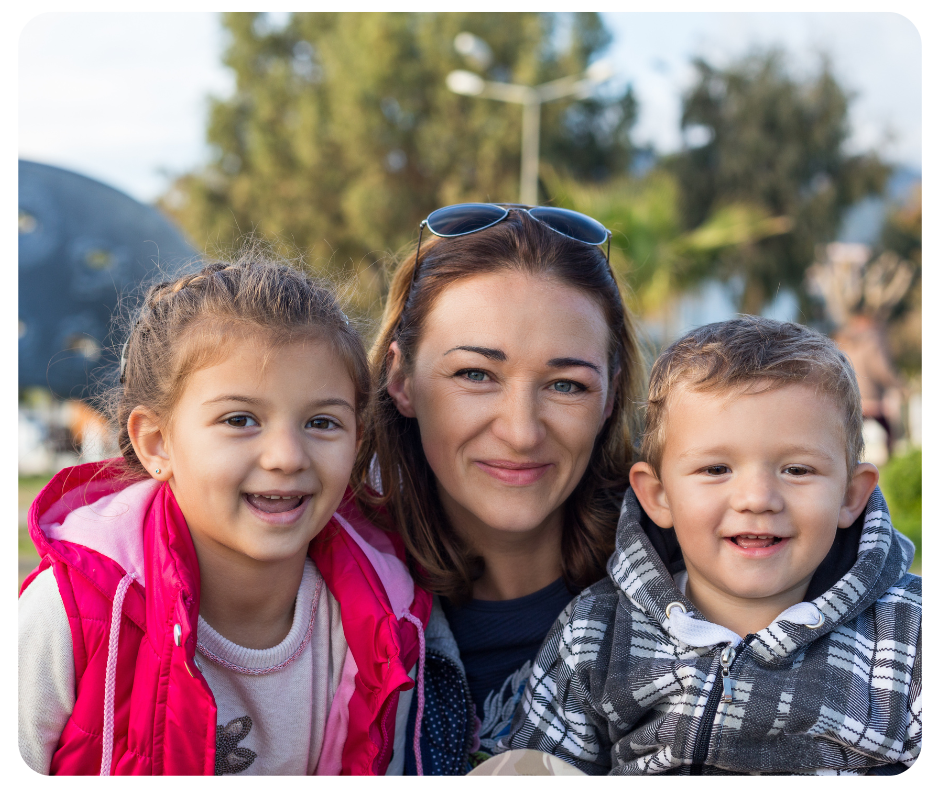 A woman is smiling with two young children, one on each side. The girl on the left is wearing a pink jacket and the boy on the right a plaid jacket. They are outdoors with trees and a structure blurred in the background.