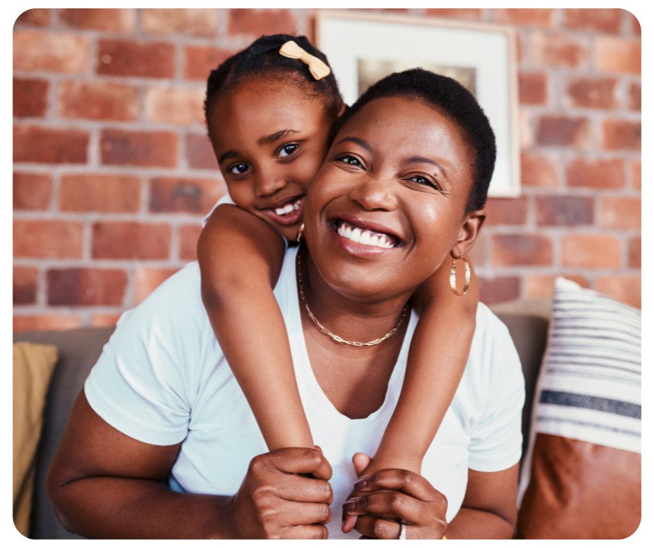 A joyful woman with short hair and hoop earrings smiles broadly while a young girl with a hair bow hugs her from behind. They are sitting on a couch against a brick wall background, and both are wearing white tops.