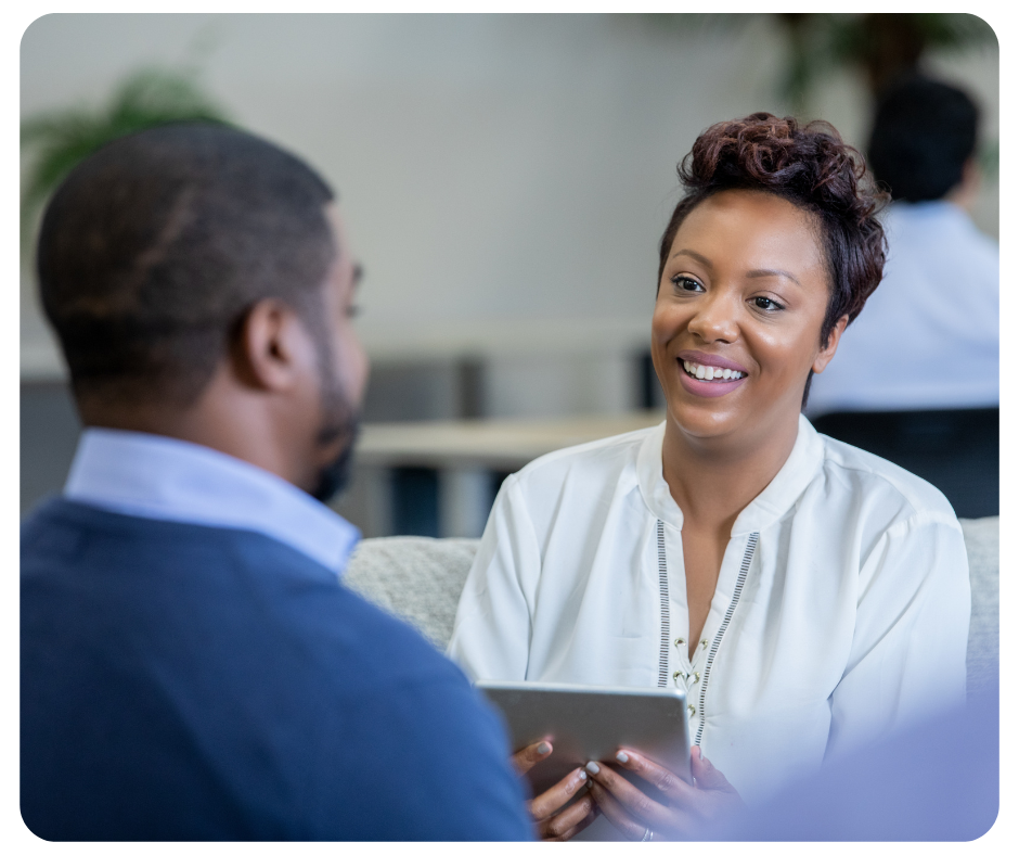 A woman smiles and holds a tablet while talking to a man in a blue sweater. They are sitting in a bright, modern office space with a blurred background.