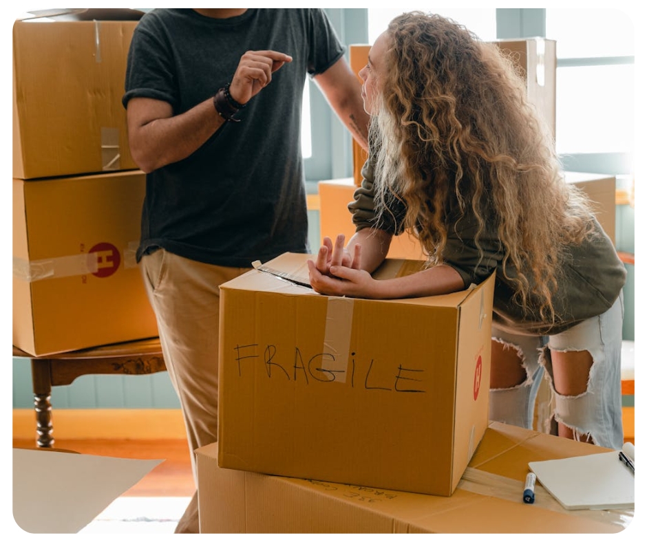 A man and a woman stand next to a stack of cardboard boxes. The woman leans on a box labeled "FRAGILE." They appear to be talking in a sunlit room. The floor is wooden, and there's packing material nearby.