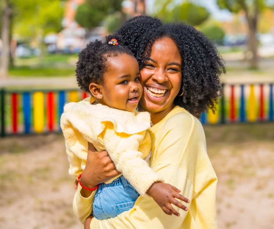 A smiling woman holds a toddler in her arms at a park. Both are wearing yellow tops, and the toddler also has blue pants. A colorful fence is visible in the background. The scene is bright and cheerful, with sunlight and greenery surrounding them.