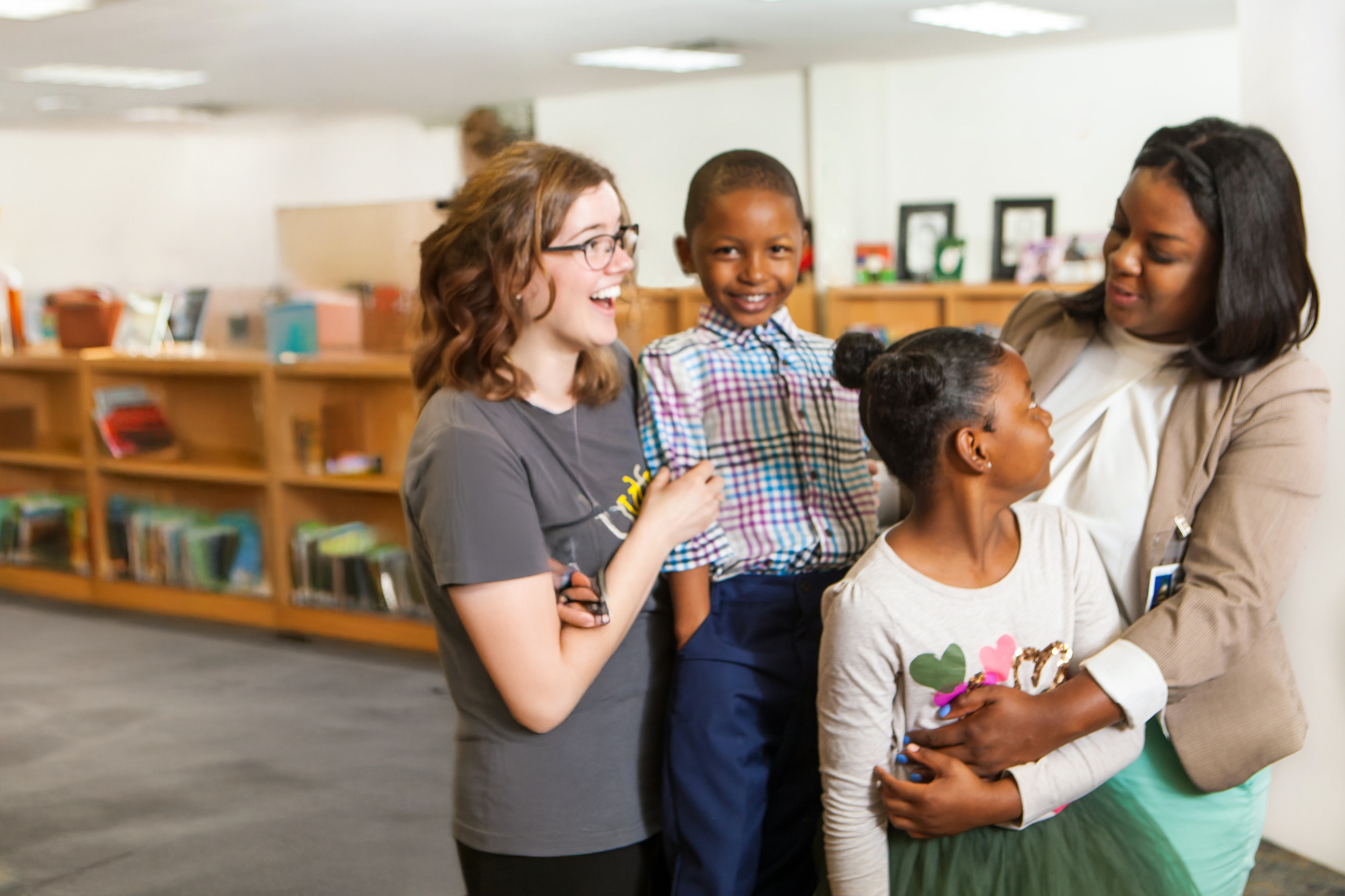 Two adults and two children are smiling and interacting in a library. Bookshelves filled with books are in the background. One adult holds a young girl, while the other adult is standing next to a young boy.