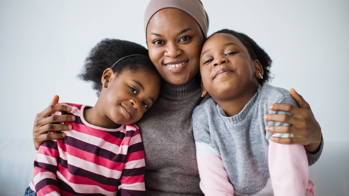 A smiling woman is seated, embracing two girls on either side. The girl on the left wears a striped shirt, and the girl on the right wears a gray sweater. They all look happy and relaxed against a plain background.