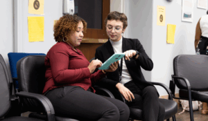 Two women sit next to each other in an office setting, looking at and discussing something on a green tablet. One gestures while the other holds the device. Empty chairs and yellow papers are visible in the background.