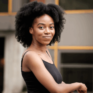 A young woman with natural curly hair, wearing a black sleeveless top, stands outdoors and smiles confidently at the camera. The background is blurred with hints of modern buildings.