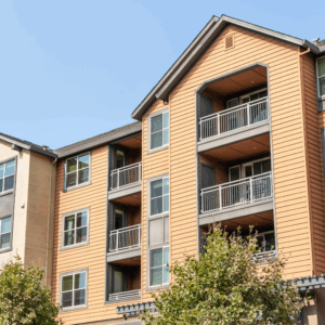 Modern apartment building with orange siding and gray trim, featuring balconies with metal railings and large windows. Green trees are in front of the building and the sky is clear and blue.