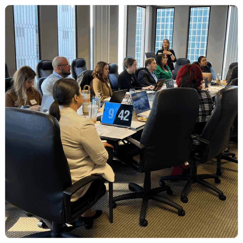 A group of people sit around a conference table with laptops and papers, listening to a woman standing and speaking near large windows in a modern office. A blue digital clock on the table displays 9:42.