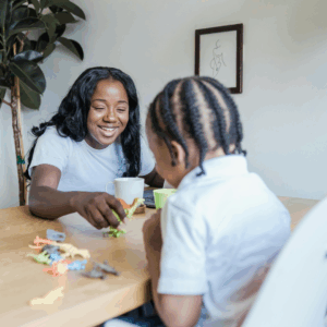 A woman and a young child play with toy animals at a wooden table. The woman is smiling, and both have dark braided hair. A potted plant and framed artwork are visible in the background.