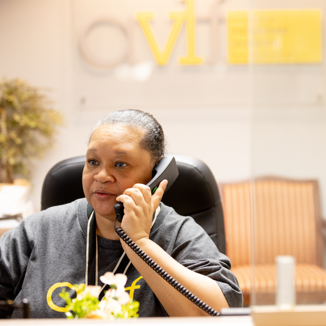 A woman wearing a gray shirt sits at a reception desk, holding a phone to her ear and smiling. A blurred office logo and seating area are visible in the background.