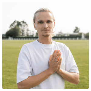 A young person with long blond hair, wearing a white t-shirt, stands outdoors on grass with hands pressed together in front of their chest, looking at the camera. Trees and a fence are in the background.
