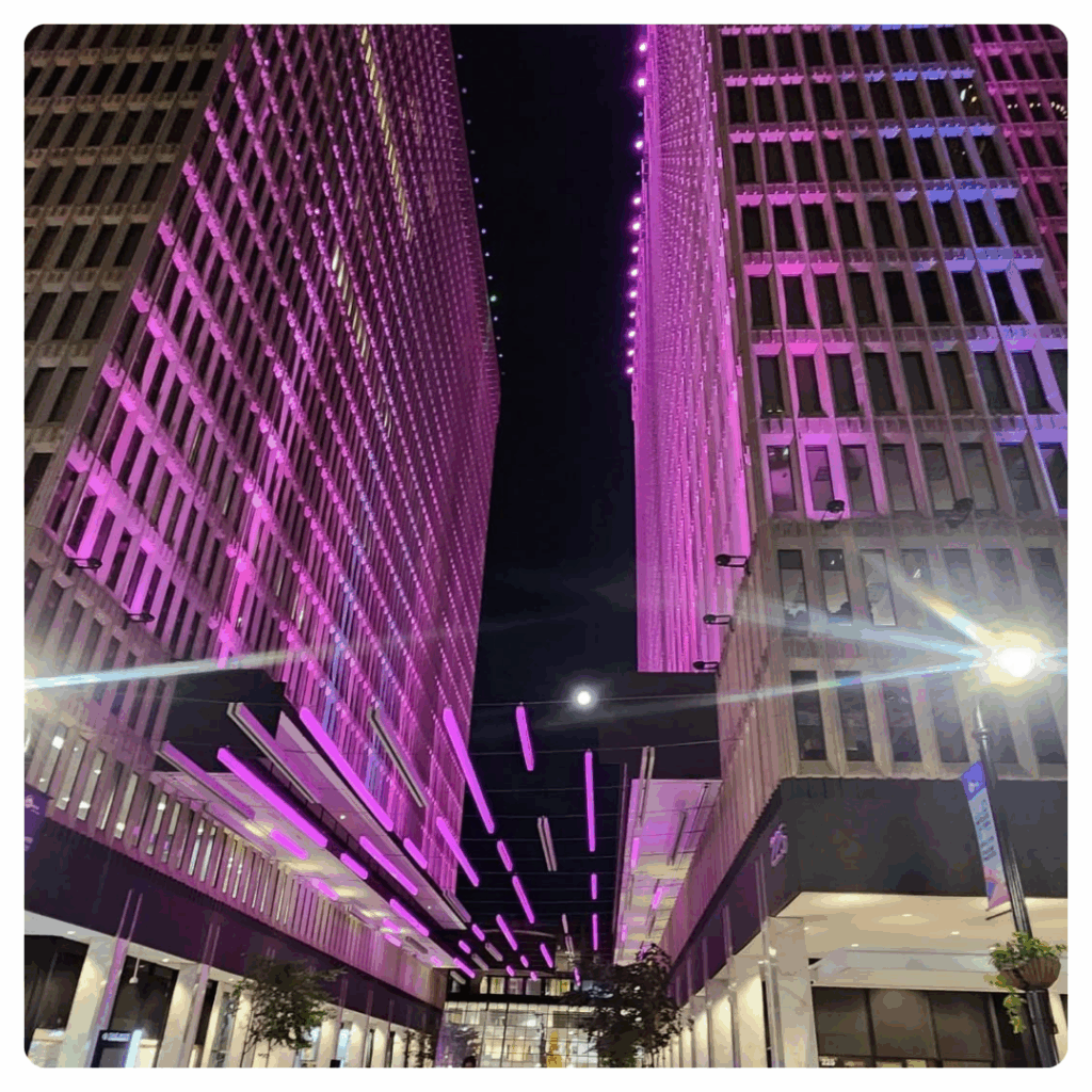 Tall buildings lit with vibrant purple and pink lights at night, with glowing horizontal light bars suspended above a walkway between the buildings, and the moon visible in the sky.