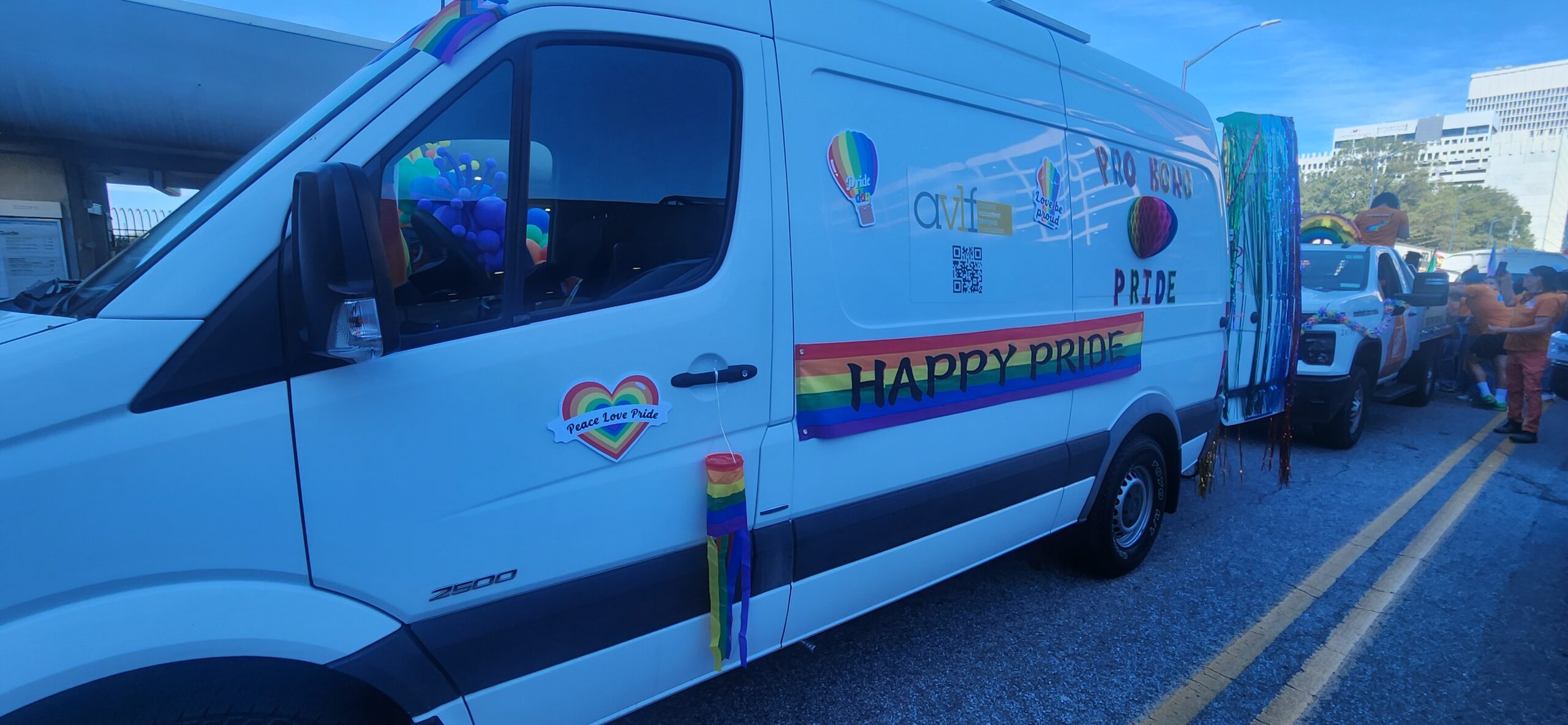 A white van decorated with rainbow flags, stickers, and a "HAPPY PRIDE" banner is parked on a street during a Pride parade. People and other decorated vehicles are visible in the background.