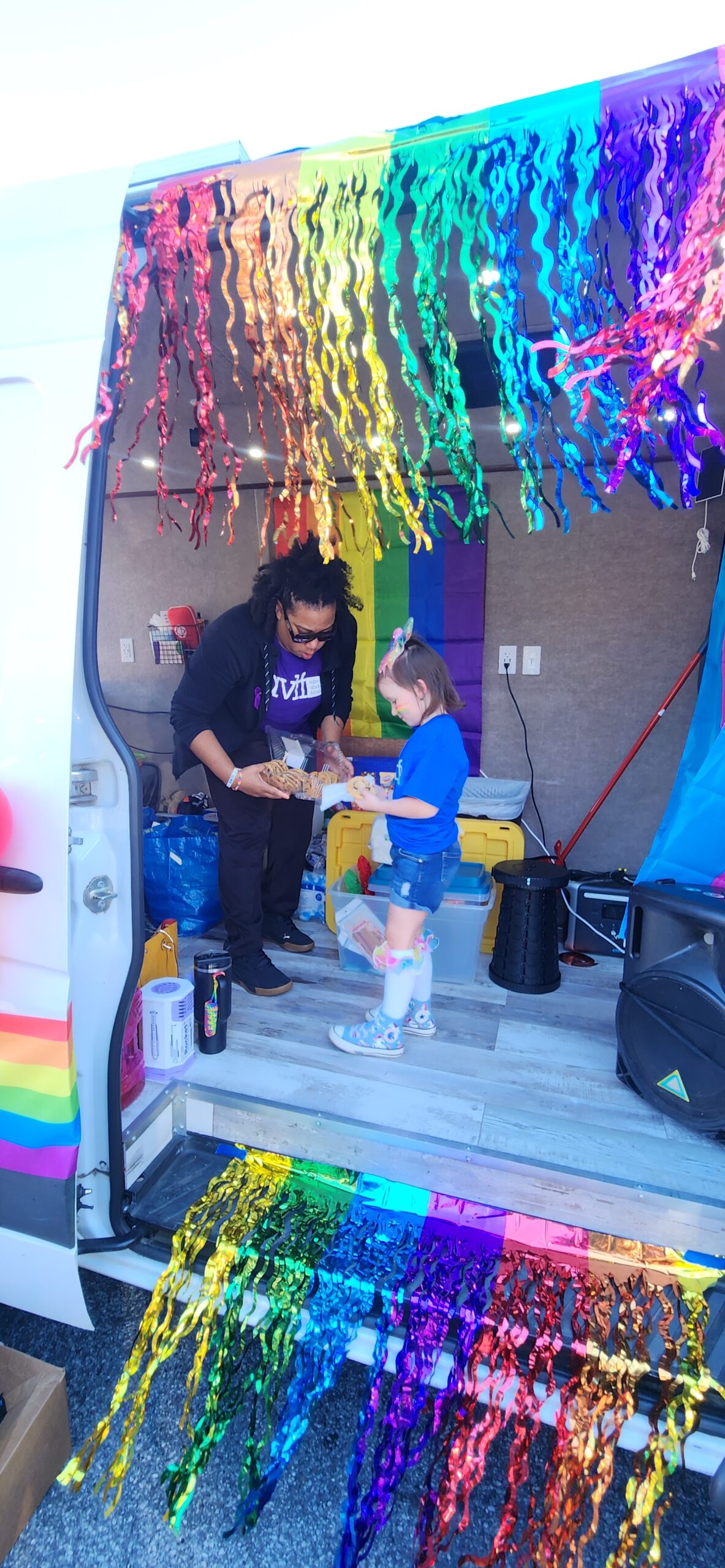 A person and a child stand inside a van decorated with rainbow streamers and Pride flags. The adult hands something to the child, who wears a unicorn headband and rainbow clothing. Sunlight brightens the festive scene.