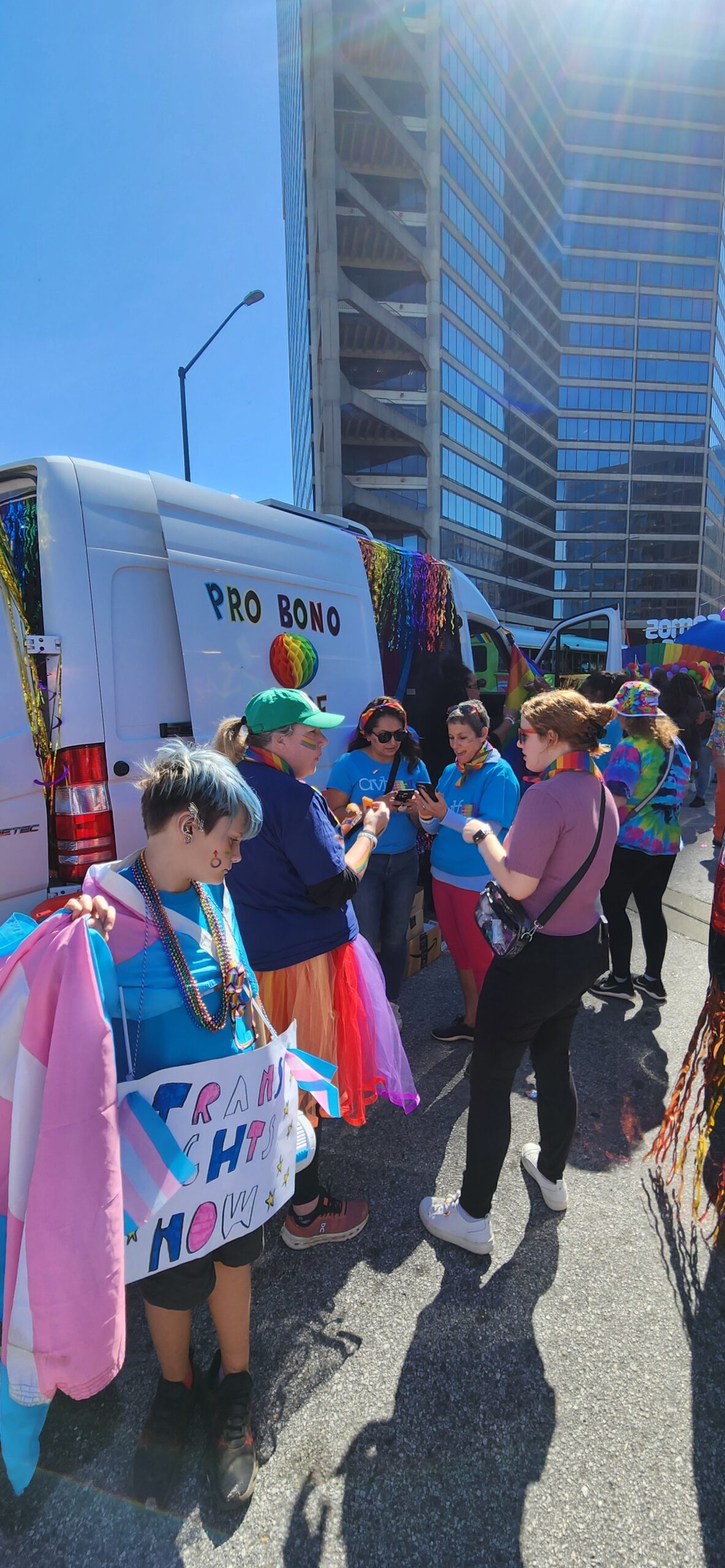 A group of people stand by a white van decorated with rainbow streamers at a street event. One person holds a “Trans Rights Now!” sign, and others wear colorful outfits and accessories. Tall buildings are visible in the background.