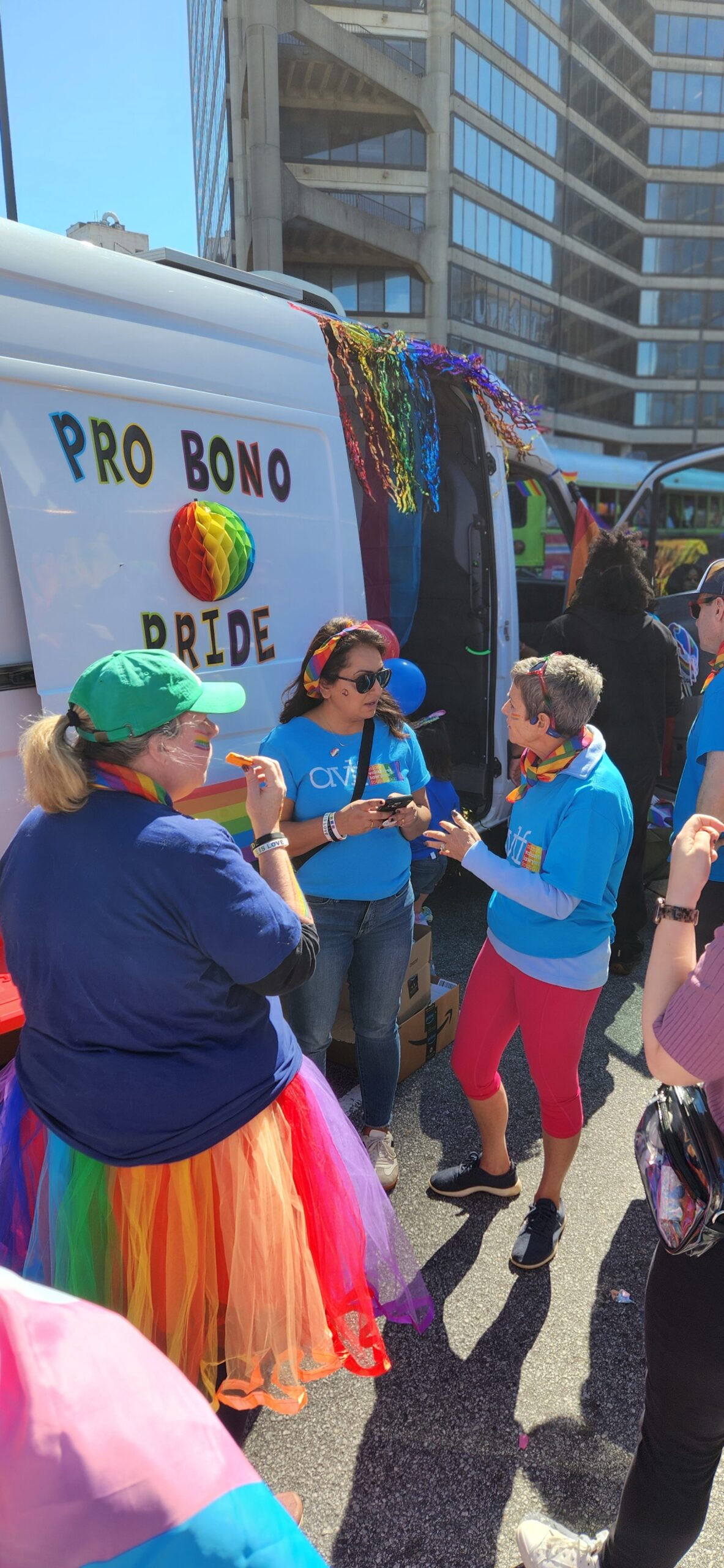 Three people in colorful outfits stand and talk in front of a van decorated with a rainbow and the words "Pro Bono Pride" during a sunny outdoor event in a city. Other participants and tall buildings are visible in the background.