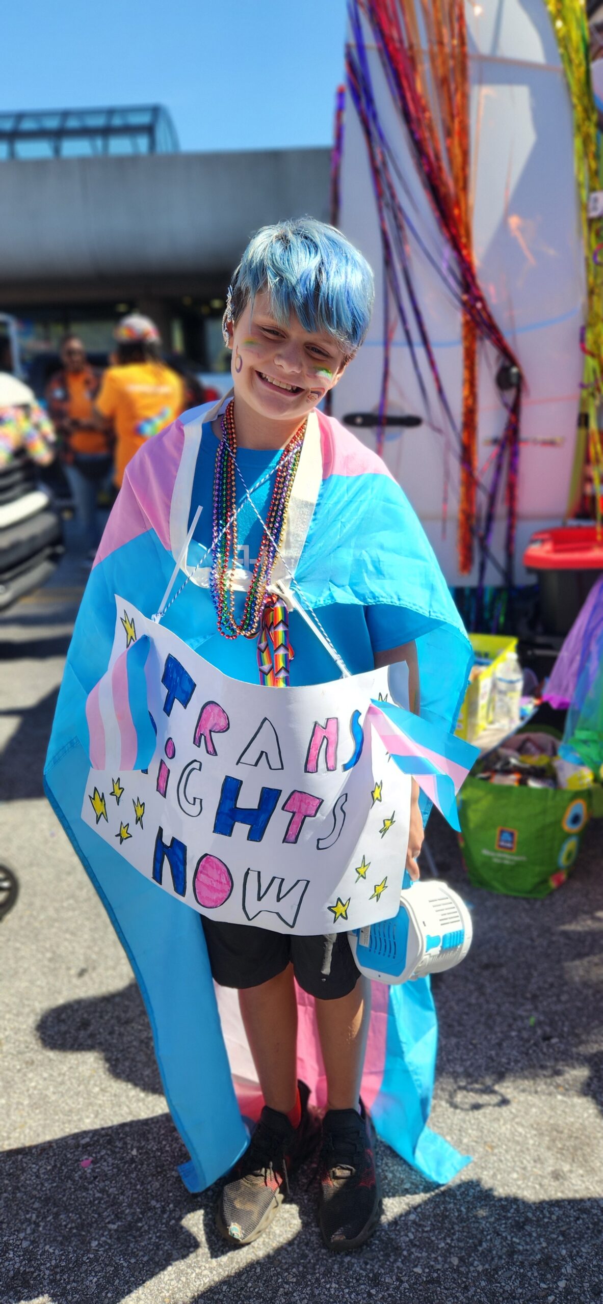 A smiling child with blue hair wears a trans pride flag as a cape, rainbow beads, and holds a colorful sign that reads "Trans Rights Now" at an outdoor event decorated with streamers.