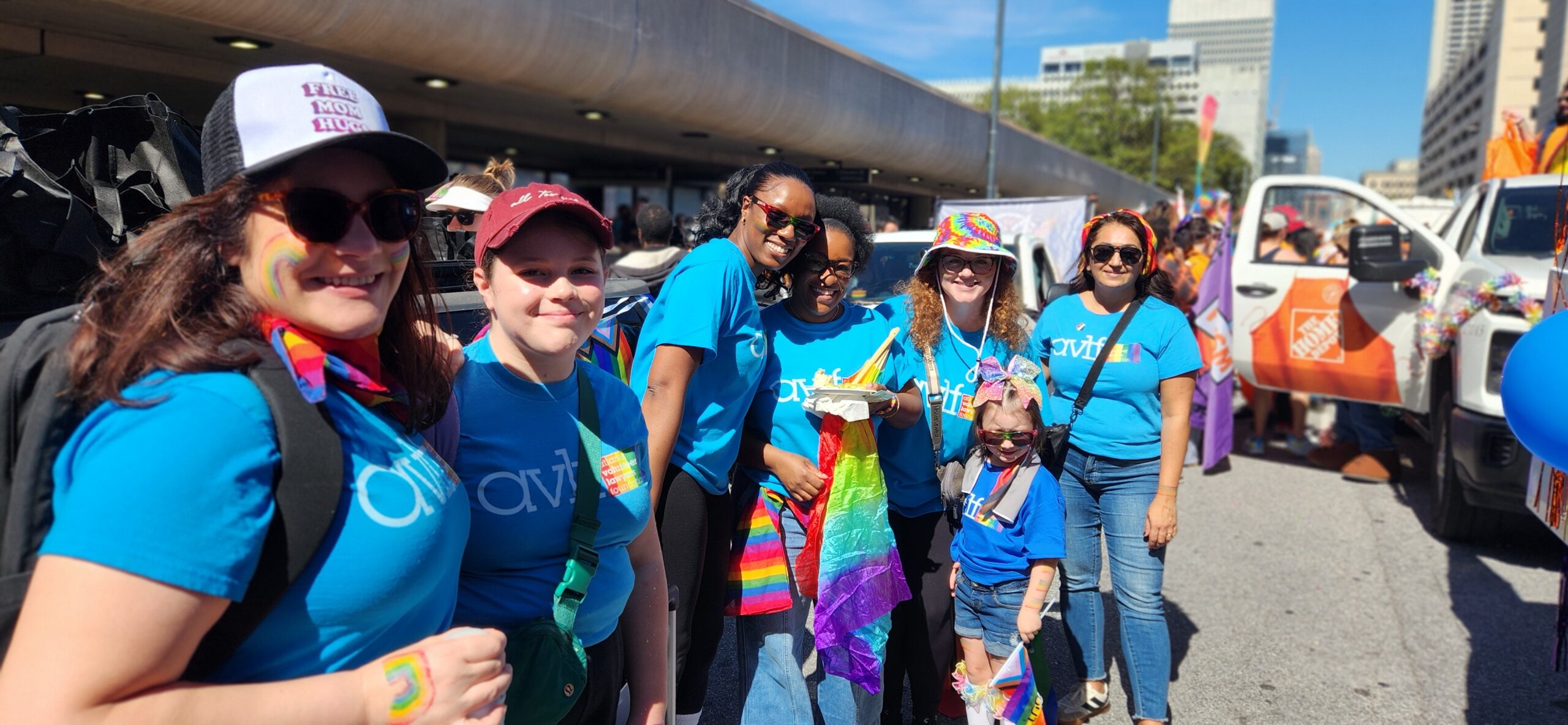 A group of people wearing blue shirts and rainbow accessories smile together at an outdoor Pride event, standing near a decorated Home Depot truck on a sunny day in the city.
