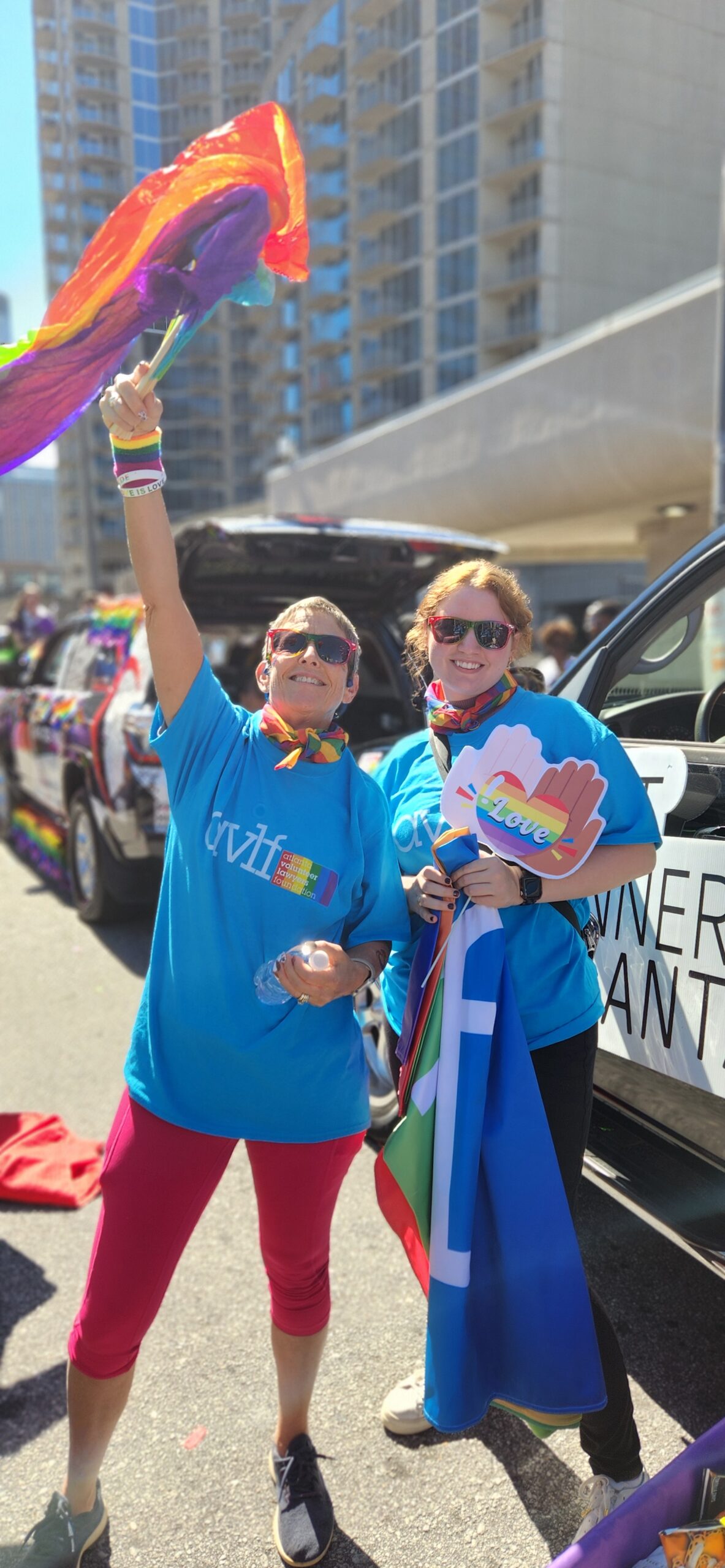 Two people wearing blue shirts, rainbow accessories, and sunglasses smile and wave rainbow flags at a pride parade. Decorated cars and tall buildings are visible in the background.