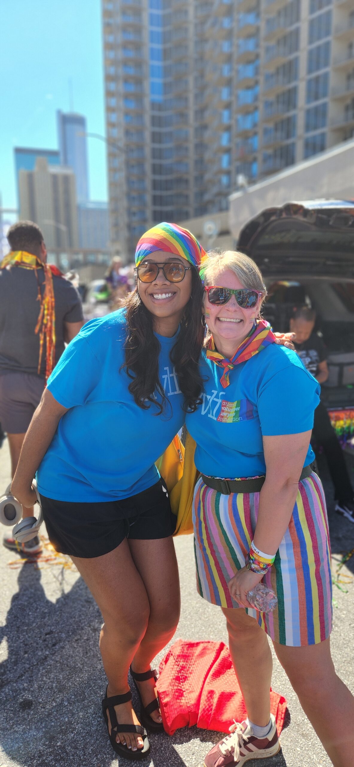 Two people wearing blue shirts and rainbow accessories smile outdoors at a parade. Tall buildings and a crowd are in the background, and both appear happy and festive.