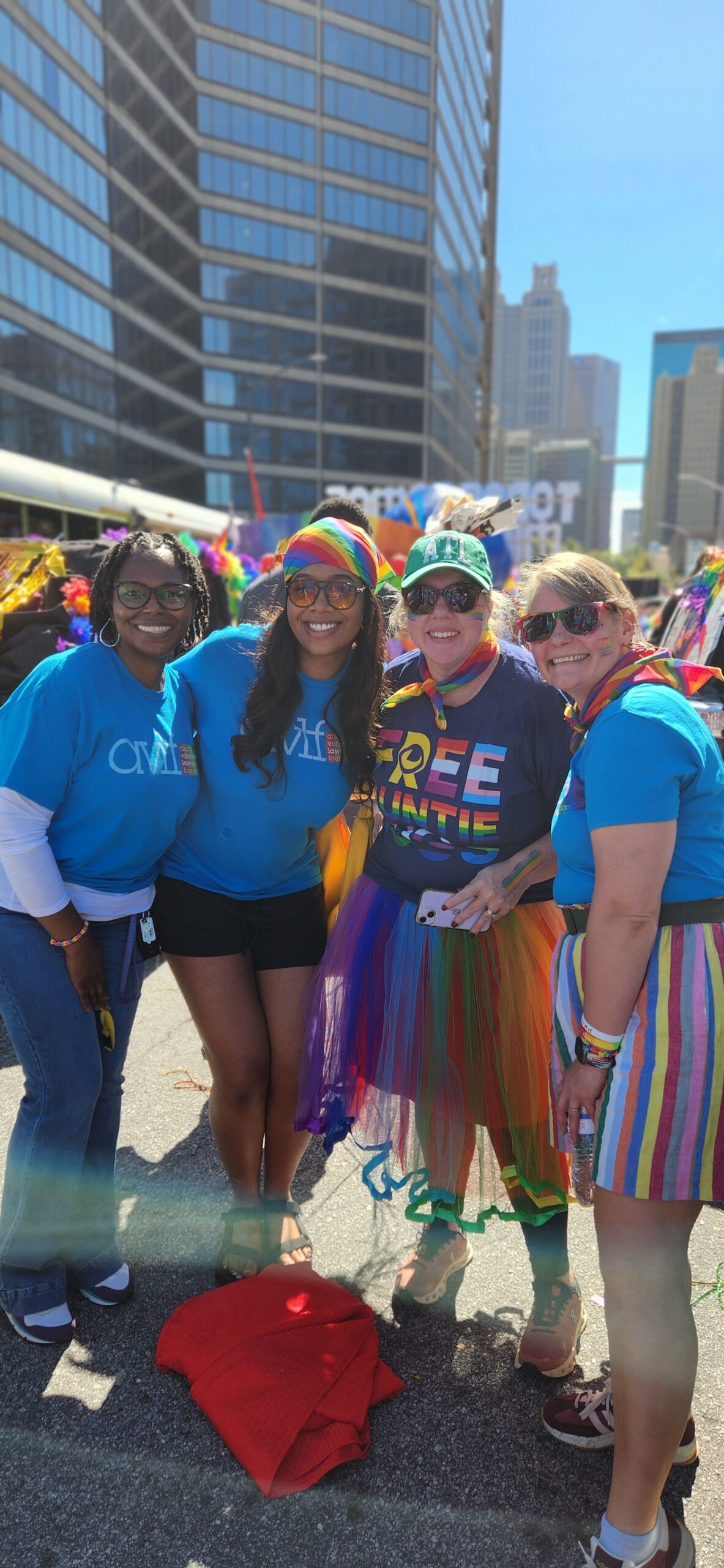 Four smiling people stand together outdoors at a Pride event, wearing colorful clothing and accessories, including rainbow skirts, hats, and shirts, with tall city buildings in the background.