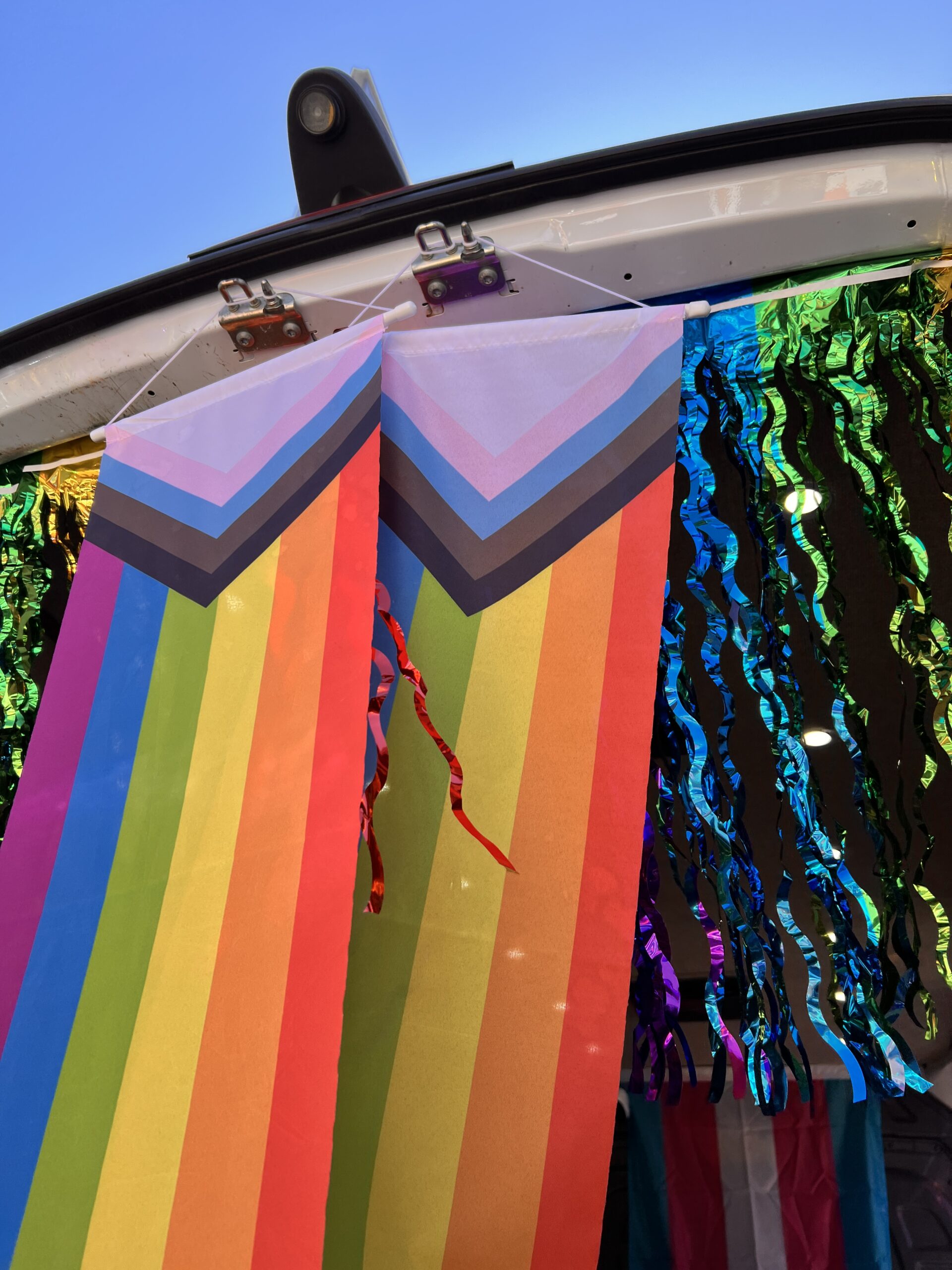 Progress Pride flags hanging from the open trunk of a car, with rainbow and black streamers in the background. The sky above is clear and blue.