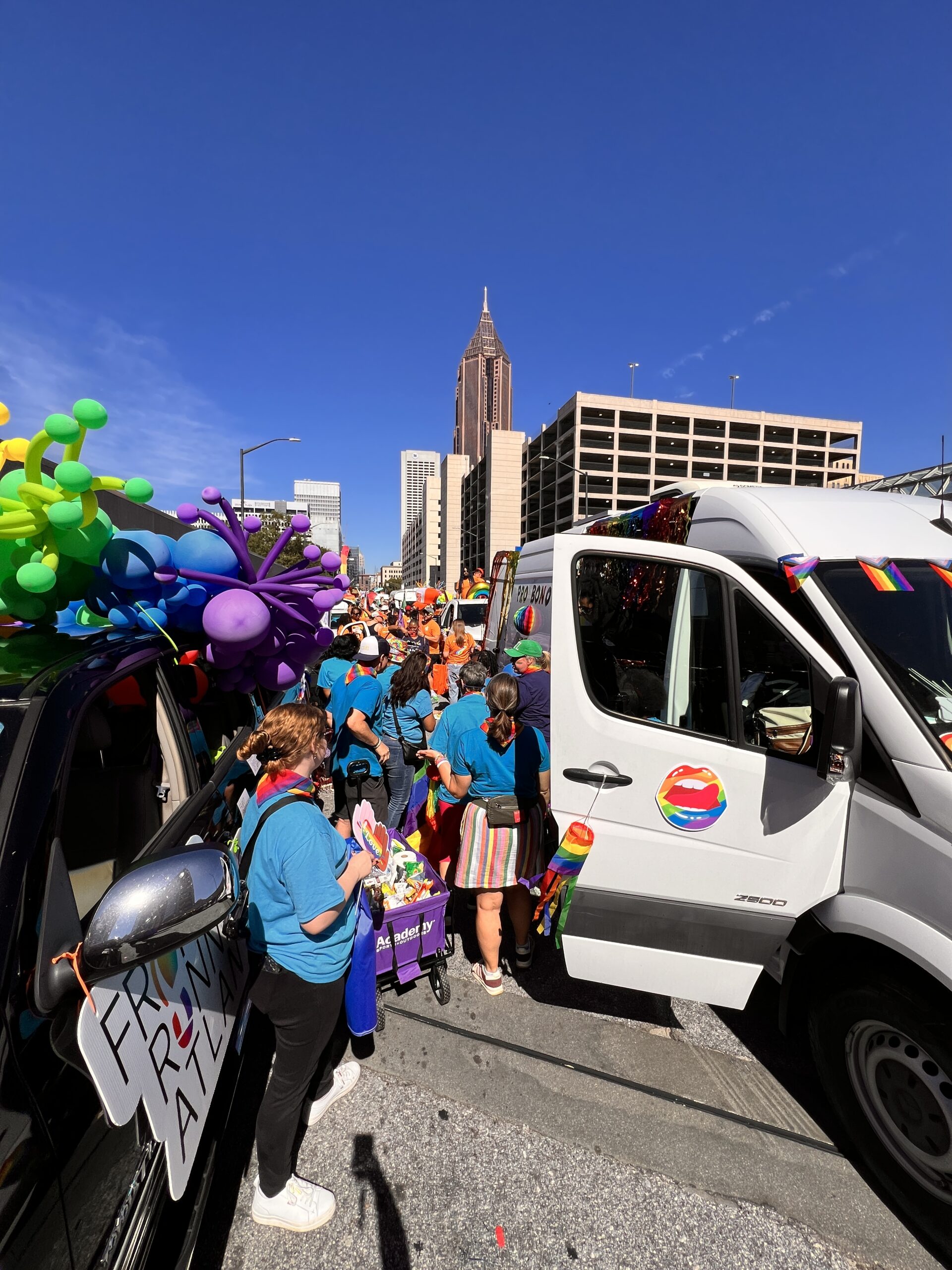 A colorful Pride parade takes place in a city street, with people in blue shirts walking between decorated vehicles, rainbow balloons, and flags. Tall buildings and a clear blue sky are visible in the background.