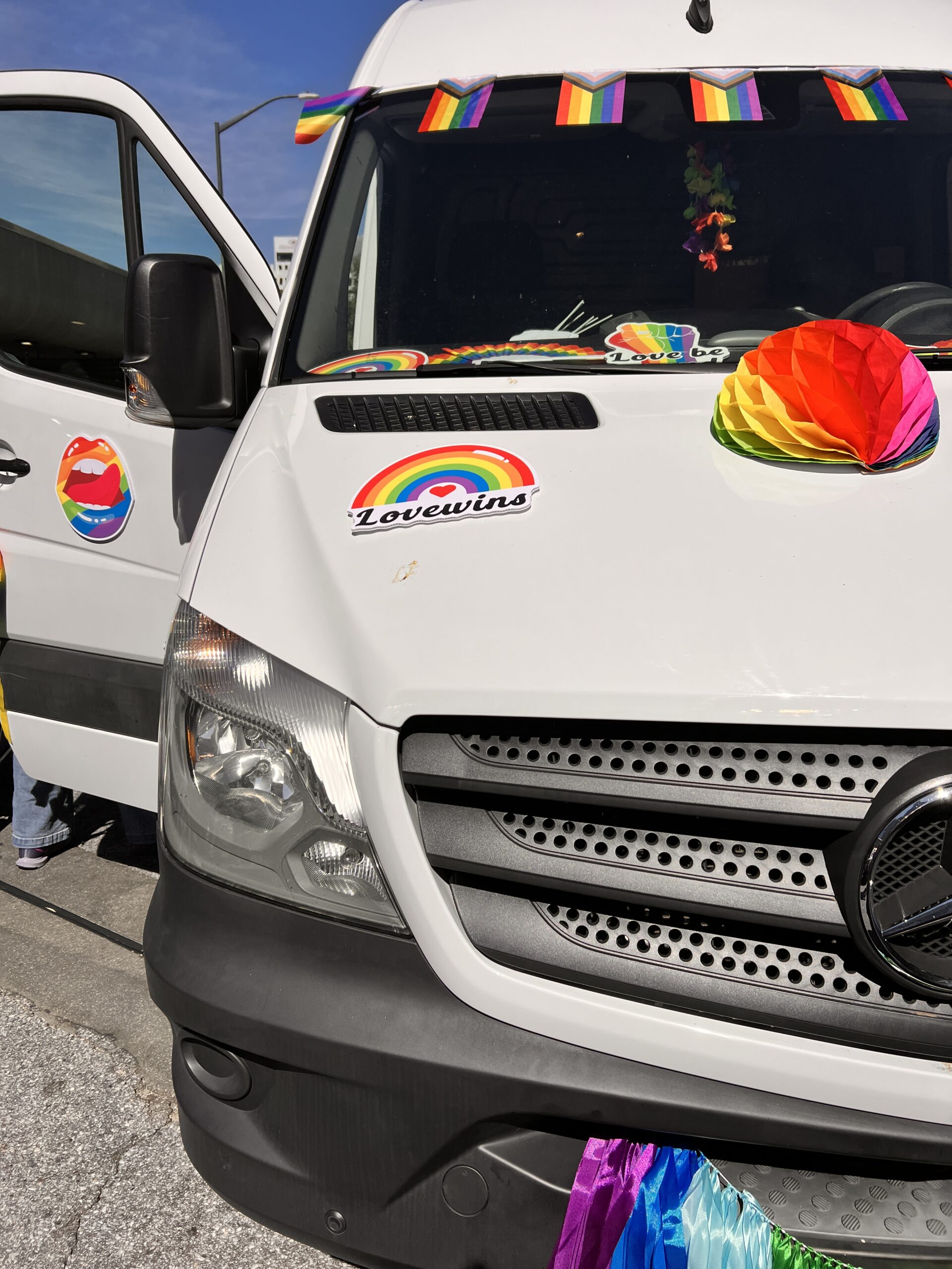 A white van decorated with rainbow flags, paper fans, and stickers, including one that says "Love wins" with a rainbow, celebrating LGBTQ+ pride. The van doors are open, and it is parked on a street.