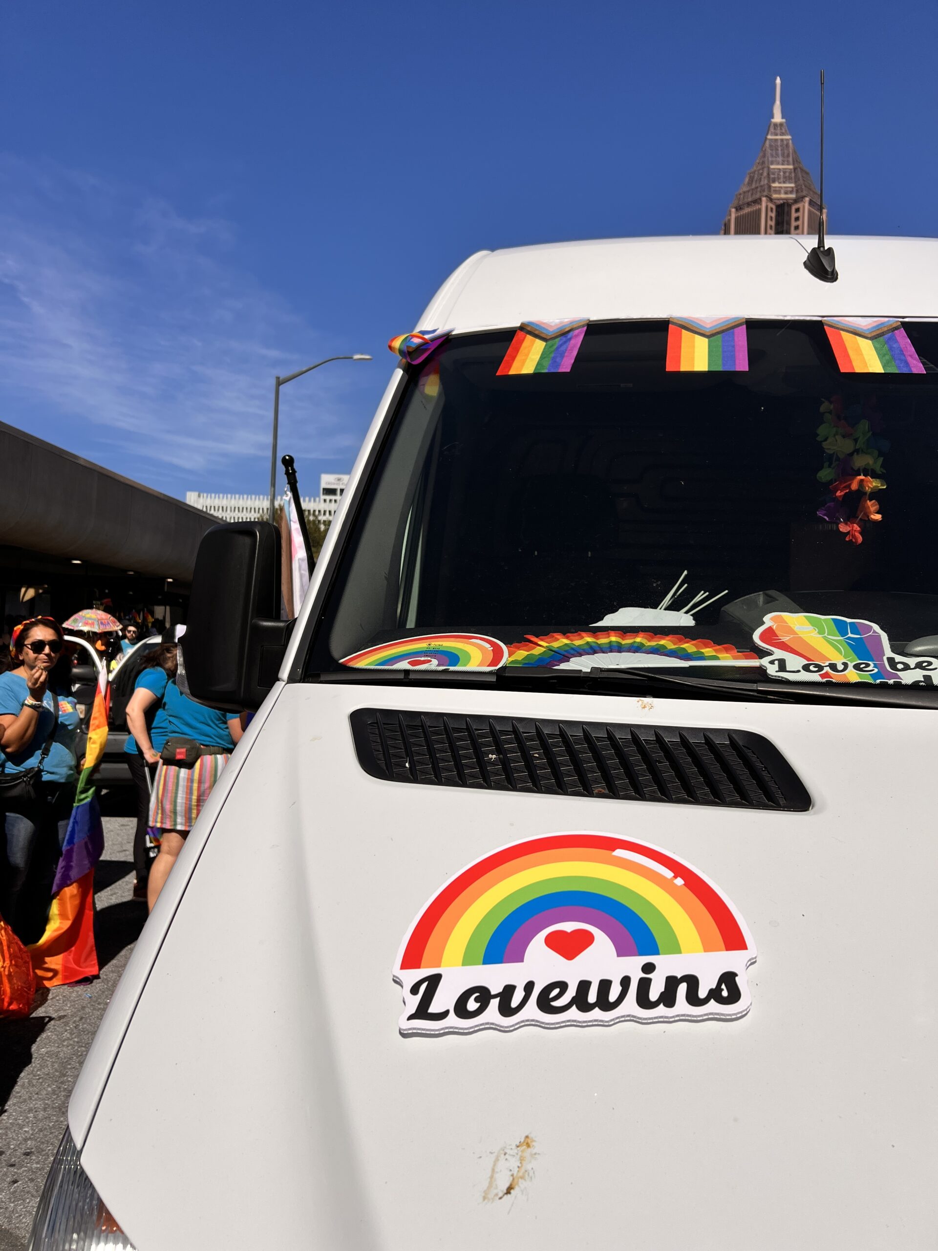 A white van decorated with rainbow flags and a sticker reading "Love wins" is parked on a sunny street during a pride event. People holding rainbow flags stand nearby under a blue sky.