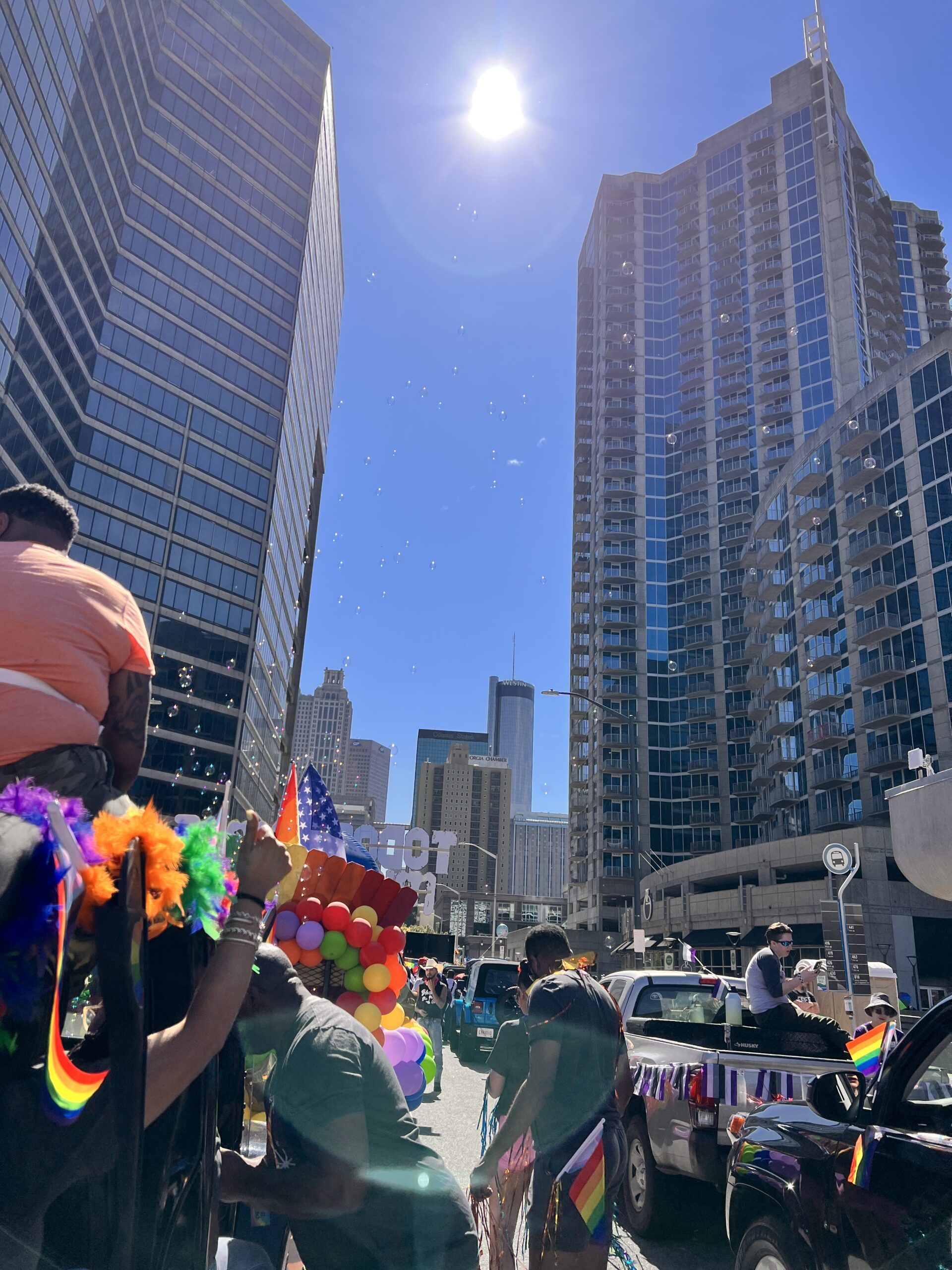A vibrant city street scene shows a Pride parade with people in colorful outfits, rainbow flags, balloons, and decorated vehicles, surrounded by tall buildings under a bright sunny sky.