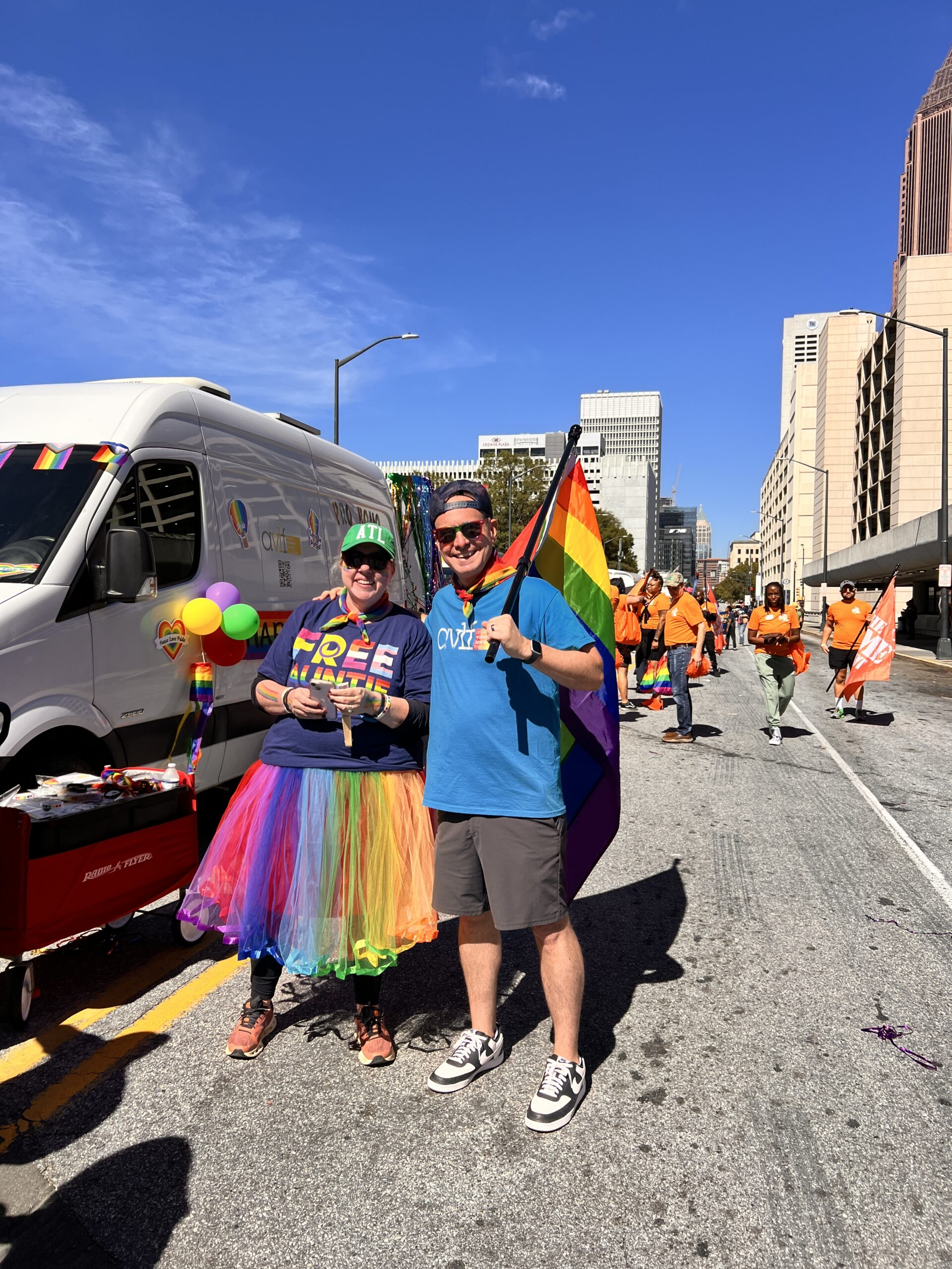 Two people smiling at a pride parade; one wears a rainbow tutu and "Free Mom Hugs" shirt, and the other holds a rainbow flag. A white van and groups in orange shirts are in the background on a sunny day.