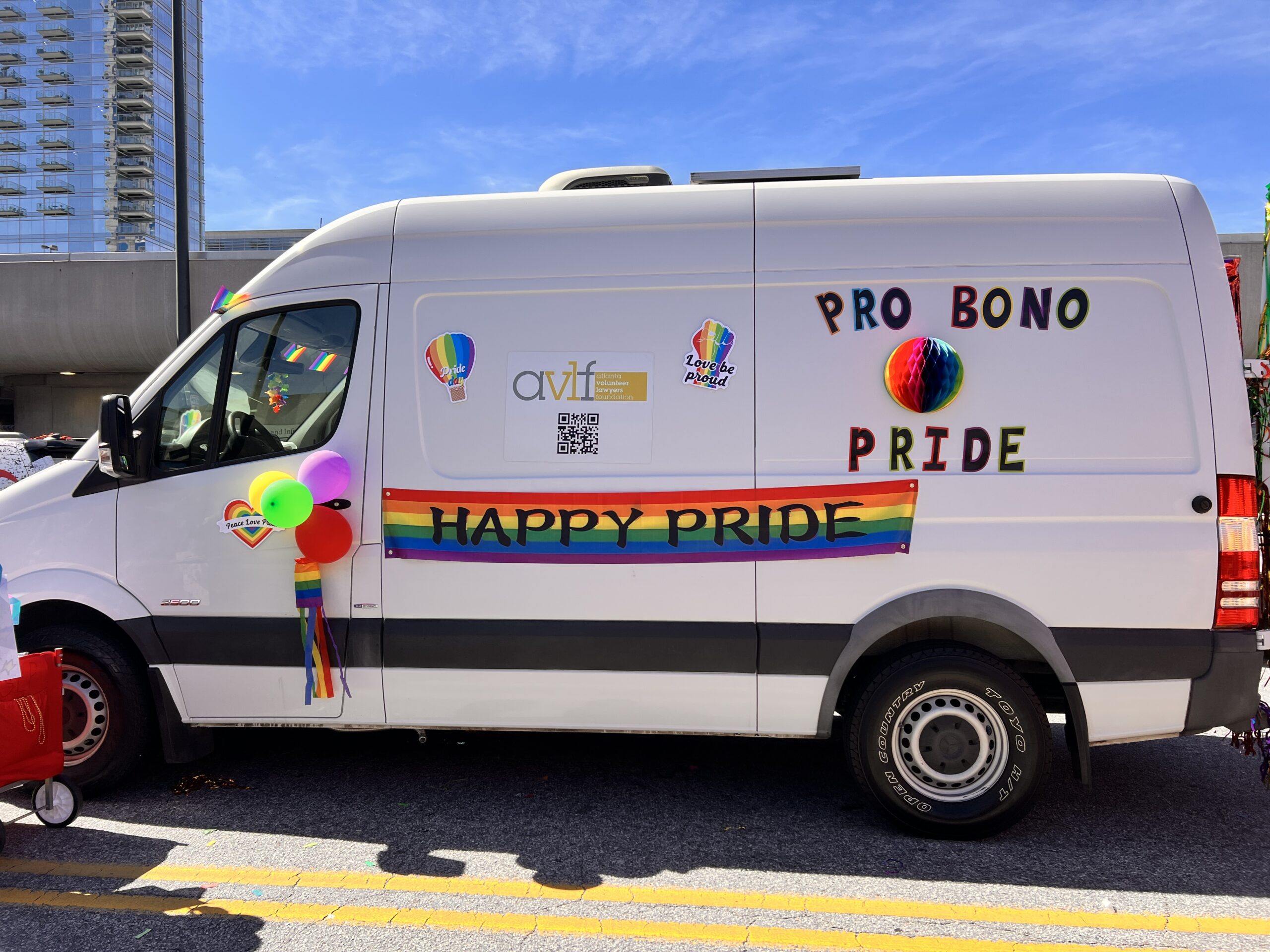 A white van decorated with rainbow balloons, a rainbow flag, and signs reading “PRO BONO PRIDE” and “HAPPY PRIDE,” participates in a parade. The background shows a tall building and a clear blue sky.
