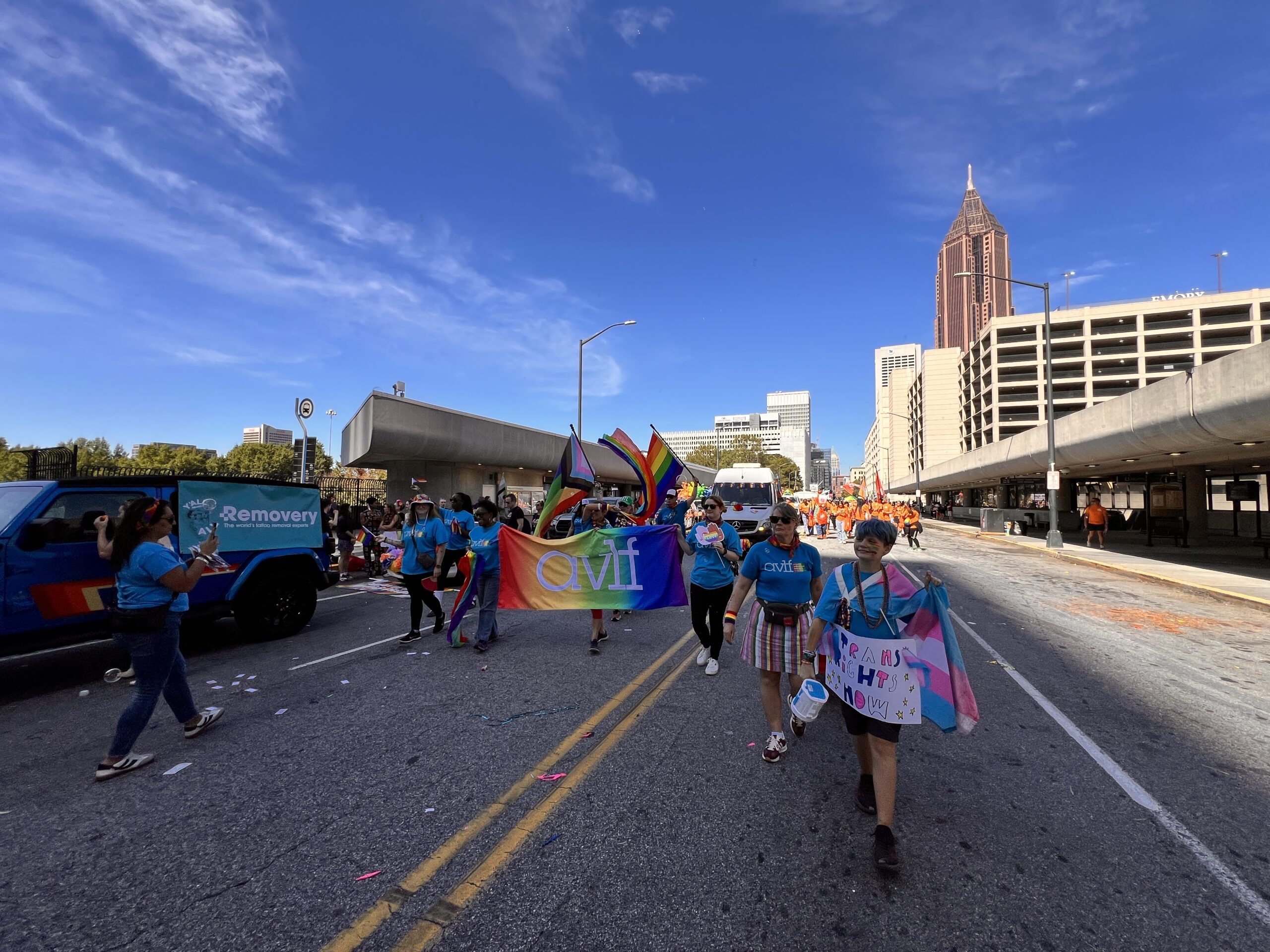 A group of people march in a Pride parade on a city street, holding colorful flags and banners under a clear blue sky, with tall buildings in the background.
