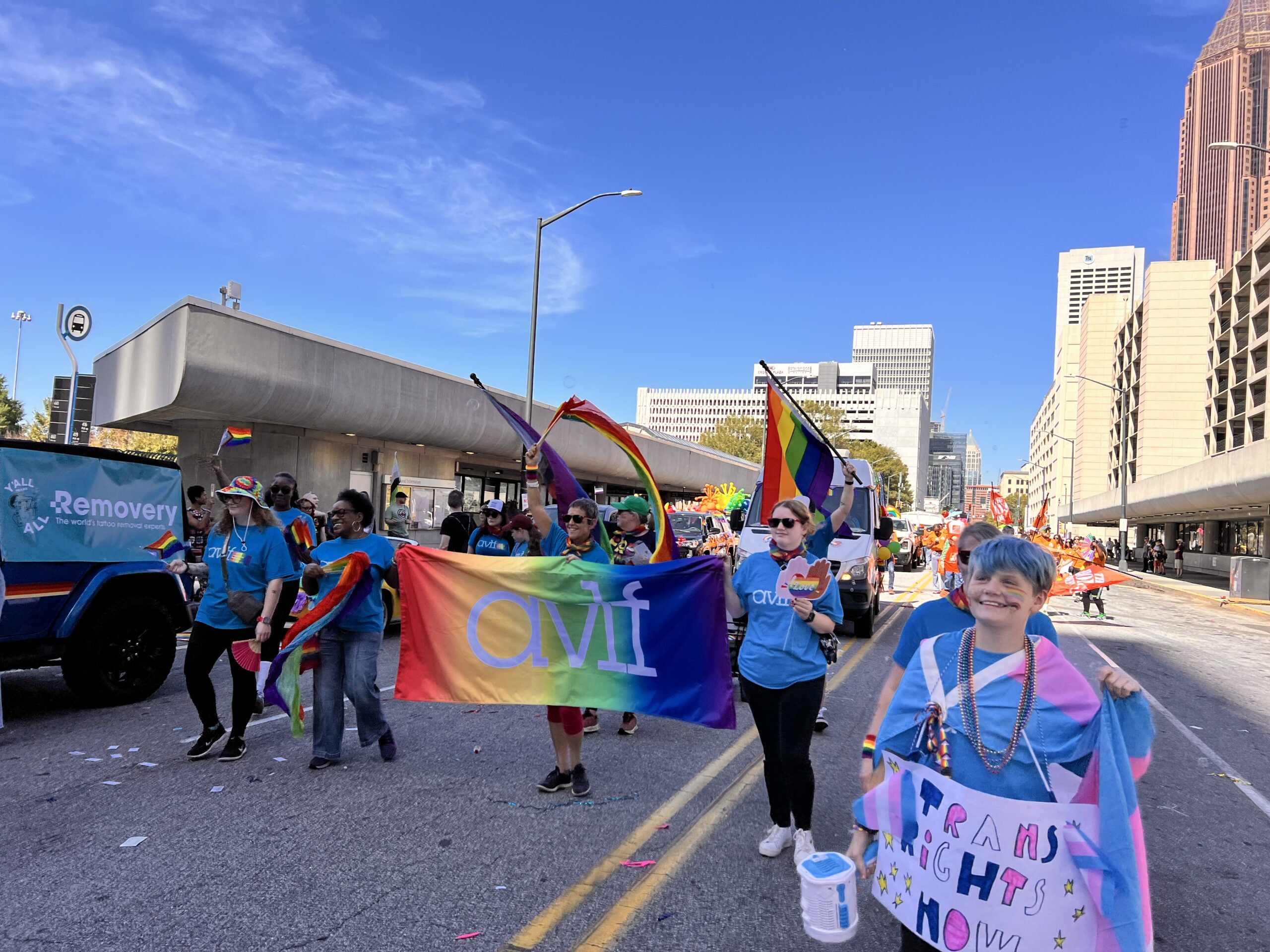 A group of people march in a city street during a Pride parade, holding rainbow flags and a colorful "avi" banner. One person in front carries a sign reading "Trans Rights Now." Skyscrapers and blue sky are visible in the background.