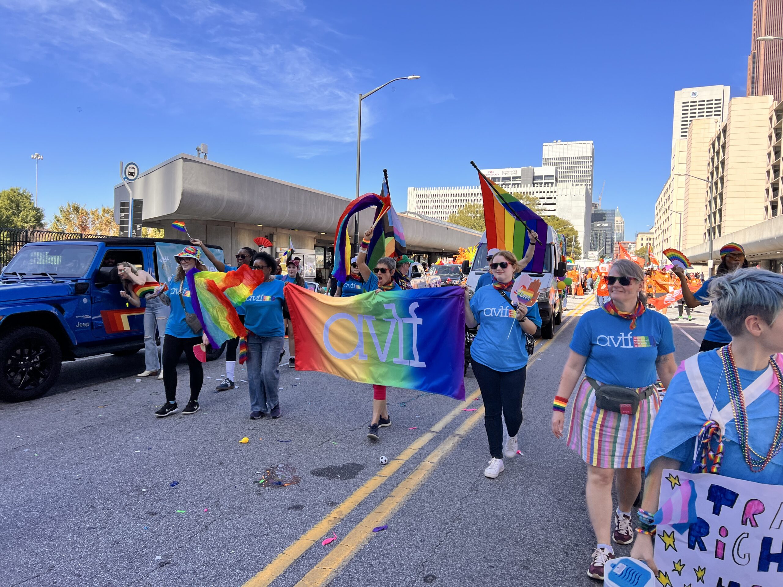 A group of people march in a pride parade, holding rainbow flags and a colorful banner that reads "avit." They wear blue shirts and walk down a city street with buildings in the background under a clear blue sky.