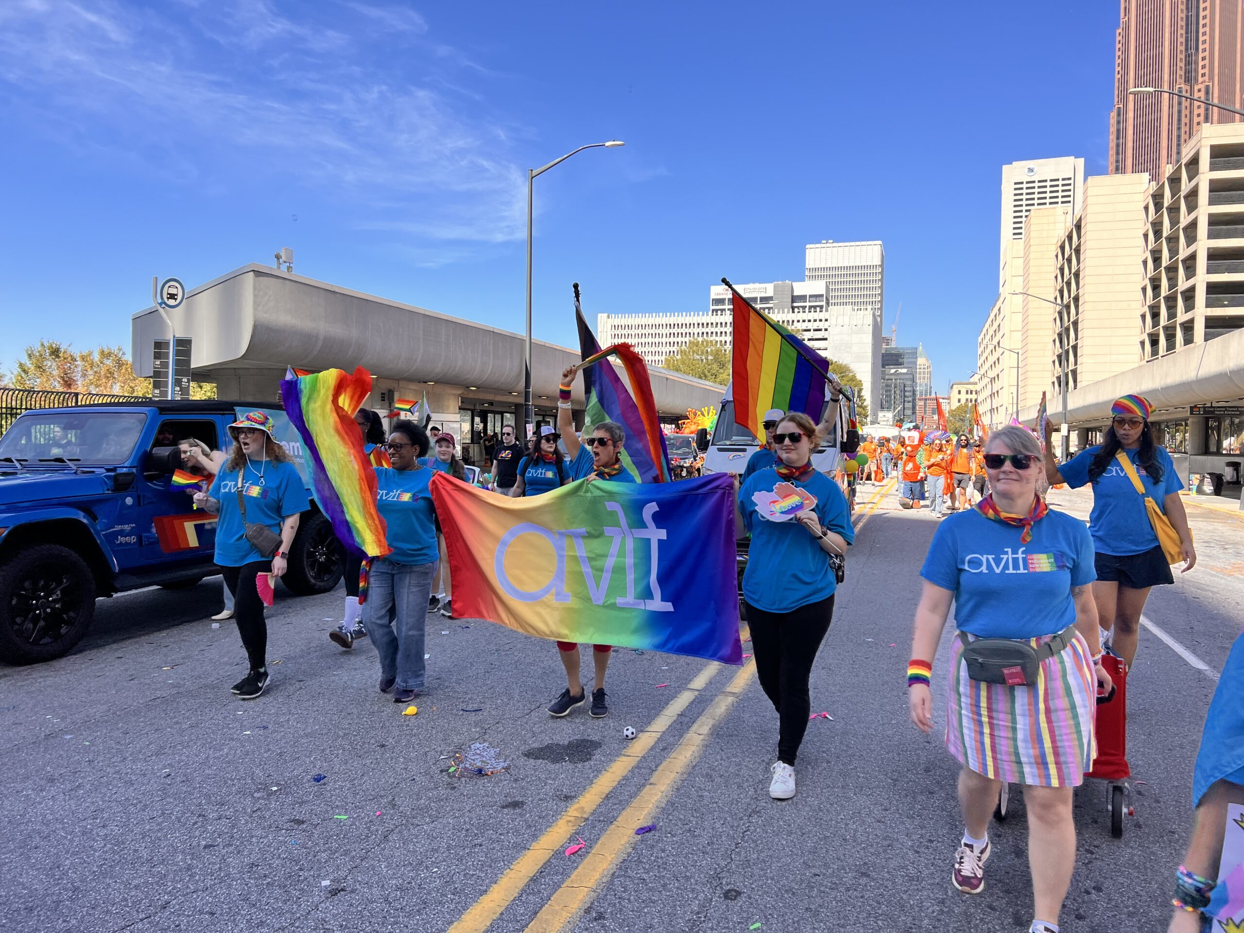 A group of people march down a city street in a pride parade, wearing blue shirts and carrying rainbow flags and a colorful banner with "avlf" on it. Tall buildings and a clear blue sky are in the background.